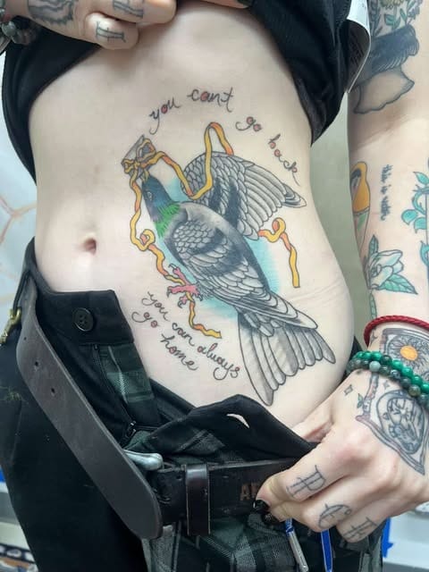a white person's stomach with a tattoo of a carrier pigeon in blues, purples, and greens, then a gray and black body. the pigeon is holding a letter in its mouth with a yellow ribbon wrapping around it. there are words around it that say "you can't go back you can always go home"